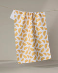 The Aubrey Fairchild Zesty Slice kitchen towel, featuring a vibrant yellow-orange citrus fruit pattern, hangs from a clothesline against a neutral background.
