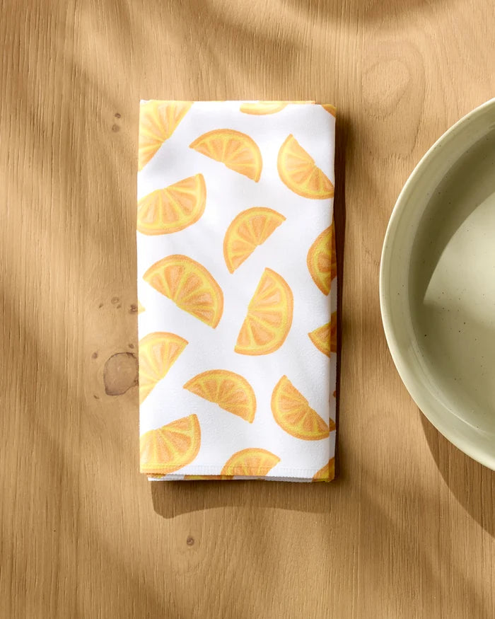 The "Zesty Slice" dinner napkin by Aubrey Fairchild, featuring a yellow lemon wedge pattern, rests folded on a light wooden table beside a beige bowl, with sunlight casting soft shadows across this cheerful table setting.