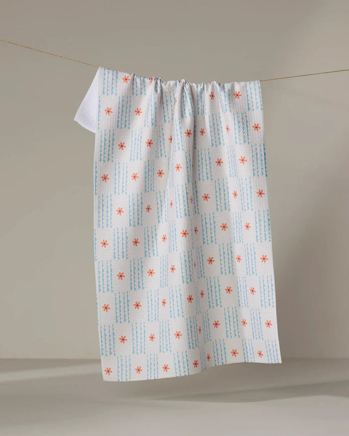 The Ziggy Floral tea towel by Elizabeth Edwards, made from recycled materials, displays blue vertical lines and red-orange asterisks and hangs from a clothesline against a neutral background.