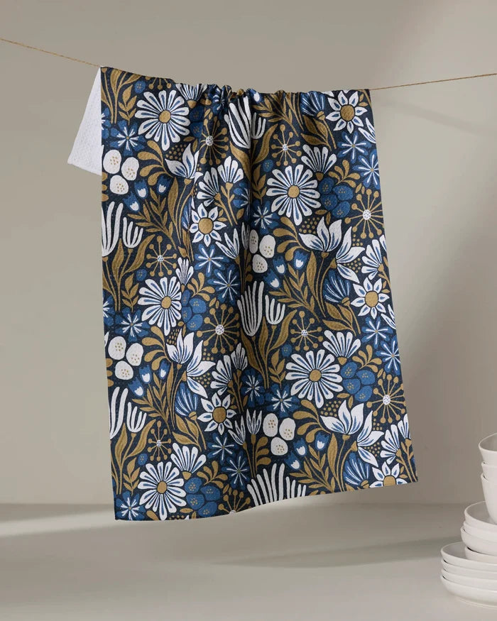 The Amy MacCready Zillia tea towel, ultra-absorbent with a botanical print in blue, white, and mustard yellow, hangs on a line; white plates are stacked in the lower right corner.