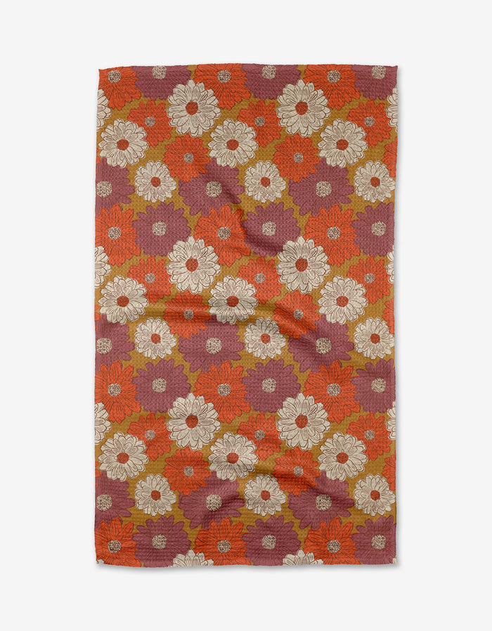 The Zinnia Forever tea towel by Kayla Ann features a retro floral design with bold white, orange, red, and olive green flowers on a reddish-purple background. Made from recycled materials, the fabric may display slight wrinkles.