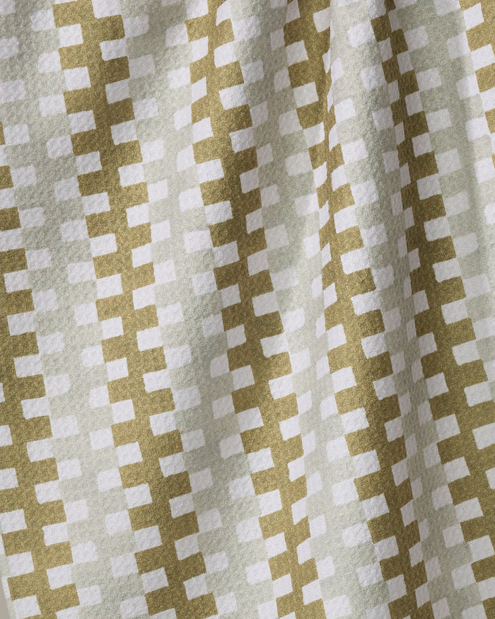 A close-up of Zippy by Studio Jeannot shows its quick-drying recycled fabric, featuring a geometric pattern of white rectangles on light green. Gentle folds and subtle shadows enhance the textured, repetitive design.