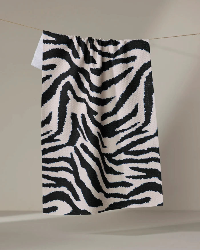 The Geometry Zira Black tea towel, featuring bold black zebra stripes and made from recycled materials, hangs from a string against a neutral background.