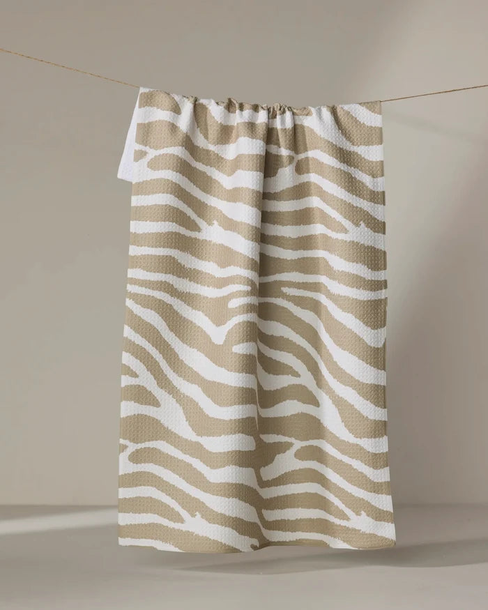 The Geometry Zira Tan tea towel, featuring beige and white zebra stripes, hangs on a thin clothesline against a softly lit neutral background.